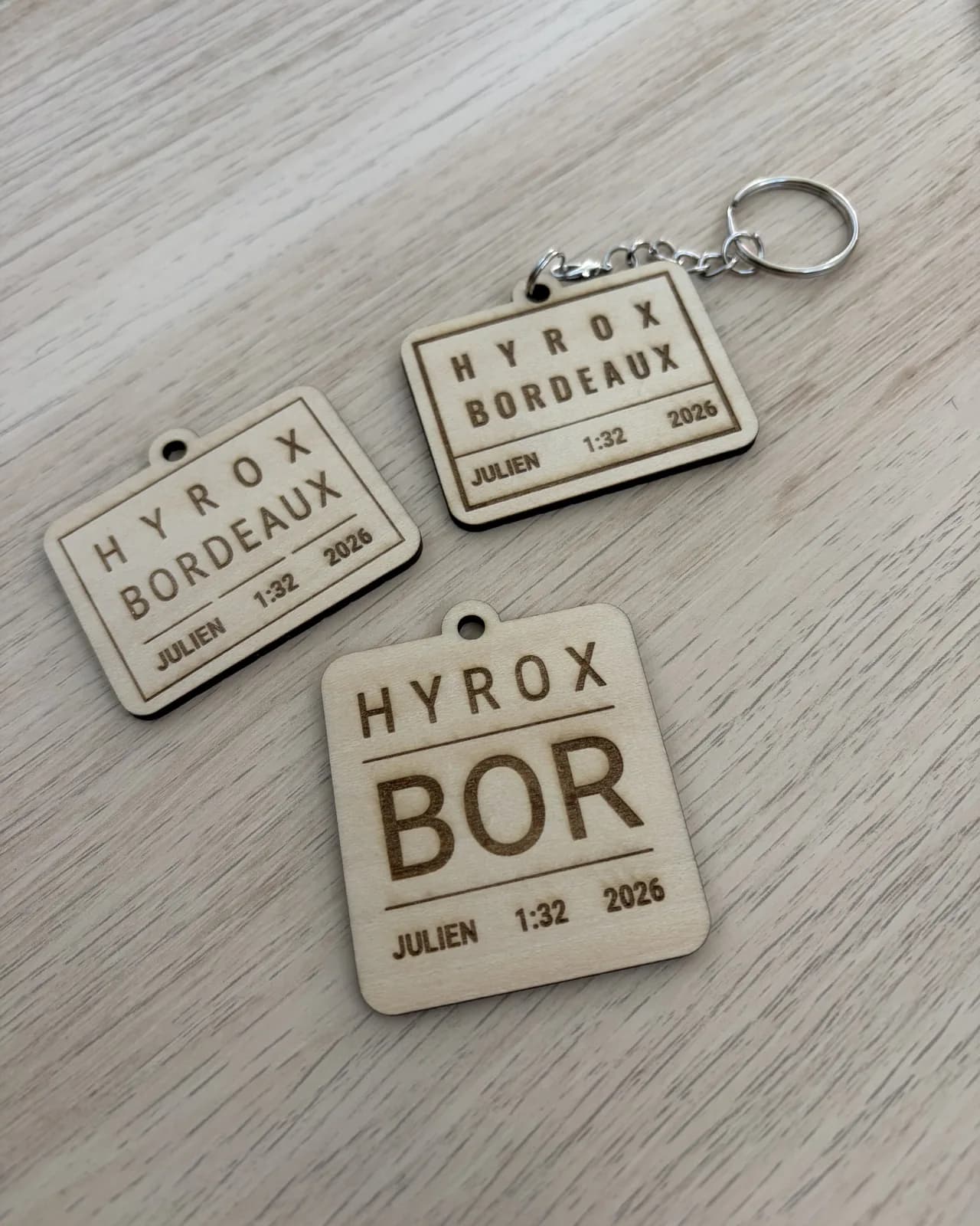 Trois exemplaires en bois clair posés sur une table : deux plaques rectangulaires, dont une montée sur un anneau à chaîne, et une plaque presque carrée à la ville abrégée en trois lettres. Chacune porte l’épreuve, la ville, un prénom, un temps et une année.