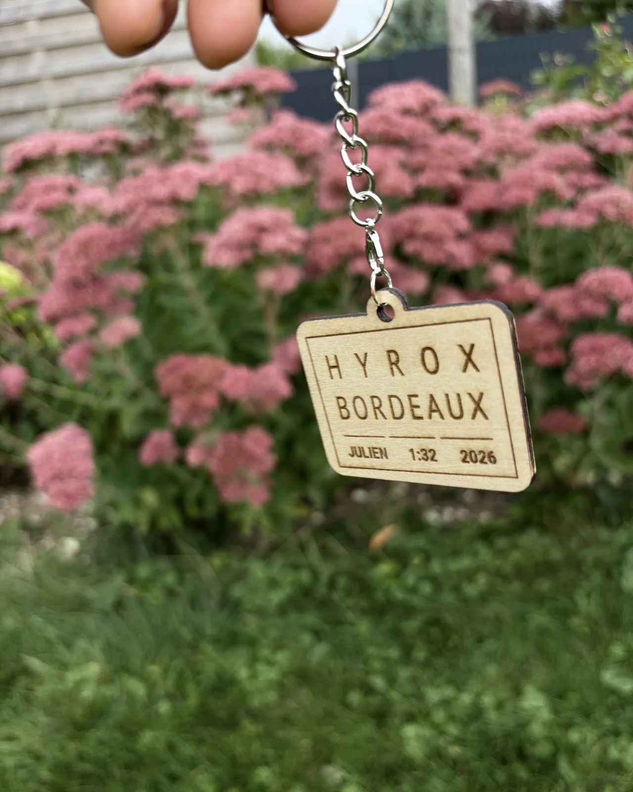 Plaque rectangulaire suspendue à un anneau à chaîne, devant un massif de fleurs roses.